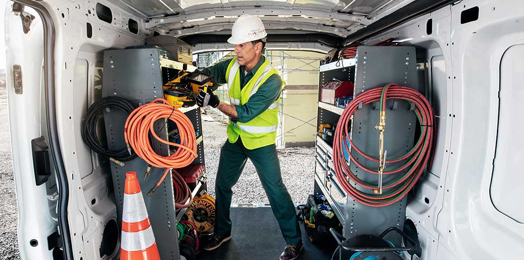 A construction worker with a hard hat retrieves equipment from the racks and bins in his 2020 Ford Transit.